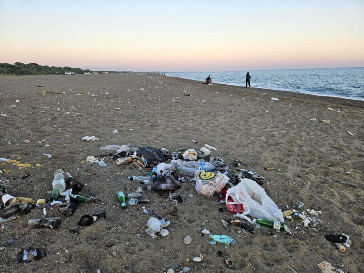Kumsaldaki manzarada, ön plandaki atık yığınlarının arkasında deniz ve gün batımı yer alırken, kirliliğin tek bir noktada kalmadığı, kumsal boyunca dağılmış olduğu dikkati çekiyor.